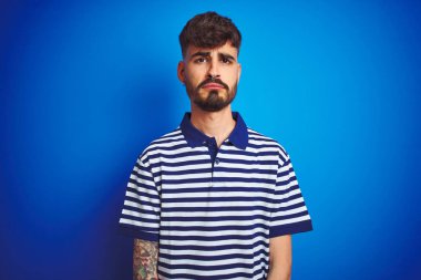 İzole edilmiş mavi arka planda duran çizgili polo giyen genç adam depresif ve endişeli, sinirli ve korkmuş. Üzücü ifade.