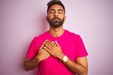 İzole edilmiş pembe arka planda duran tişört giyen genç Hintli adam elleri göğsünde, gözleri kapalı ve yüzünde minnet dolu bir jest ile gülümsüyor. Sağlık kavramı.
