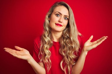 Kırmızı izole edilmiş arka plan üzerinde duran basit tişört giyen genç ve güzel bir kadın. Kolları ve elleri havada, kafası karışmış bir ifade. Şüphe kavramı.