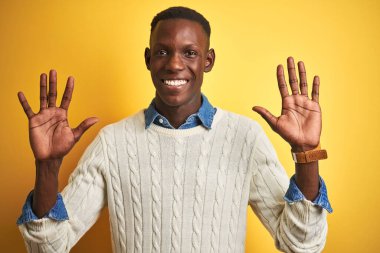İzole edilmiş sarı arka planda kot gömlek ve beyaz süveter giyen Afro-Amerikalı adam on numarayı gösterirken kendinden emin ve mutlu bir şekilde gülümsüyor..