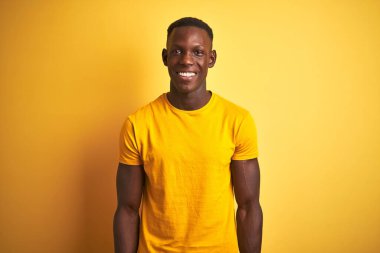 Yüzünde mutlu ve havalı bir gülümsemeyle izole edilmiş sarı arka planda dikilen sıradan tişörtler giyen Afro-Amerikan genç adam. Şanslı kişi..
