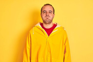 Yağmurluk giyen genç adam izole edilmiş sarı arka planda duruyor komik yüzlü yanakları şişiriyor. Havayla şişmiş ağız, çılgın bir ifade..
