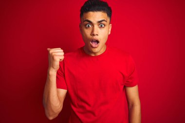 İzole edilmiş kırmızı arka plan üzerinde duran Brezilyalı genç adam şaşırmış bir şekilde elini kenarı işaret ediyor, ağzı açık şaşkın bir şekilde..