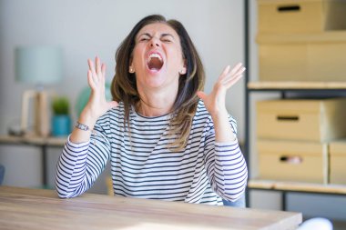 Orta yaş kıdemli kadın evde masada oturan çılgın ve deli çığlık ve agresif ifade ve kollar ile bağırdı kaldırdı. Hayal kırıklığı kavramı.