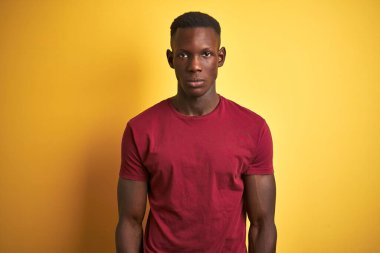 Kırmızı tişört giyen genç Afro-Amerikalı adam izole edilmiş sarı arka planda duruyor. Yüzünde ciddi bir ifadeyle rahatlamış. Basit ve doğal bir şekilde kameraya bakıyorsun..
