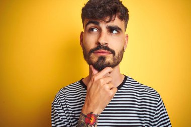 Dövmesi olan genç adam çizgili t-shirt giyen çene üzerinde el ile izole sarı arka plan üzerinde duran soru, dalgın ifade düşünme. Düşünceli bir yüzle gülümsüyor. Şüphe kavramı.