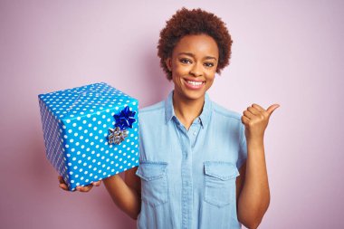 Afrika kökenli Amerikalı bir kadın pembe izole edilmiş arka planda doğum günü hediyesini tutuyor ve gülümseyen bir gülümsemeyle baş parmağıyla işaret ediyor.