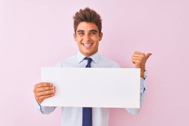 Genç yakışıklı işadamı izole pembe arka plan üzerinde duran afiş tutan işaret ve mutlu yüzü gülümseyerek yan başparmak ile gösteren
