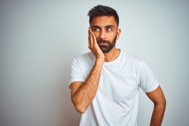 İzole edilmiş beyaz arka planda duran tişört giyen genç Hintli adam kollarını kavuşturarak depresyondan sıkılmış ve yorgun görünüyor..