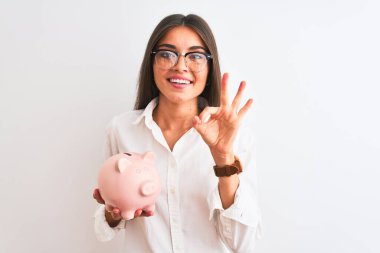 Gözlüklü güzel bir iş kadını kumbarasını izole edilmiş beyaz arka planda tutuyor tamam işareti yapıyor, mükemmel bir sembol.