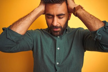 Yeşil tişörtlü genç Hintli adam izole edilmiş sarı arka planda duruyor. Baş ağrısı, acı ve migren yüzünden çaresiz ve stresli. Ellerini başının üstüne koy..