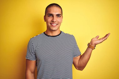 Sarı izole edilmiş arka plan üzerinde çizgili tişört giyen genç yakışıklı adam mutlu bir şekilde sunum yapıyor ve avuç içi kameraya bakıyor..