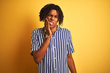 Rastgele çizgili tişört giyen Afro bir adam izole edilmiş sarı arka plan üzerinde diş ağrısı ya da diş ağrısı yüzünden acı dolu bir ifadeyle ağıza dokunuyor. Dişçi konsepti.