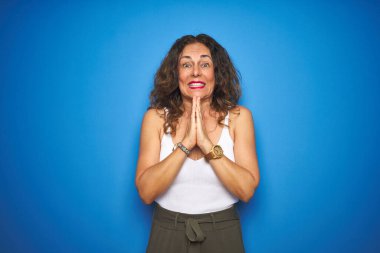 Middle age senior woman with curly hair standing over blue isolated background praying with hands together asking for forgiveness smiling confident. clipart
