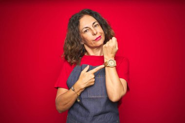 Kırmızı izole edilmiş arka planda önlük giyen orta yaşlı bir kadın. Aceleyle saati, sabırsızlığı ve rahat bir ifadeyle kameraya bakıyor.