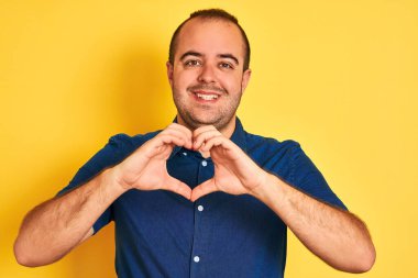 İzole edilmiş sarı arka planda dikilmiş kot gömlekli genç bir adam. Aşkla gülümsüyor. Elleriyle kalp sembolü ve şekli gösteriyor. Romantik konsept.