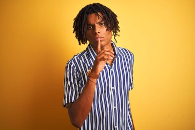 Rasta desenli tişörtlü Afro bir adam izole edilmiş sarı arka planda sessiz olmayı diliyor. Sessizlik ve gizli konsept.