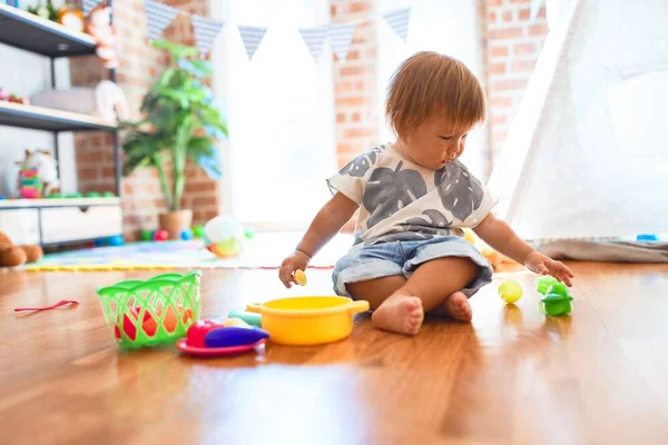 幼児は床に座ってプラスチック製のカトラリーや食べ物を使って食事をしています幼稚園のおもちゃの多くの周りに — ストック写真