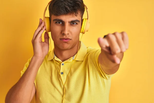 Young indian man listening to music using headphones over isolated yellow background pointing with finger to the camera and to you, hand sign, positive and confident gesture from the front