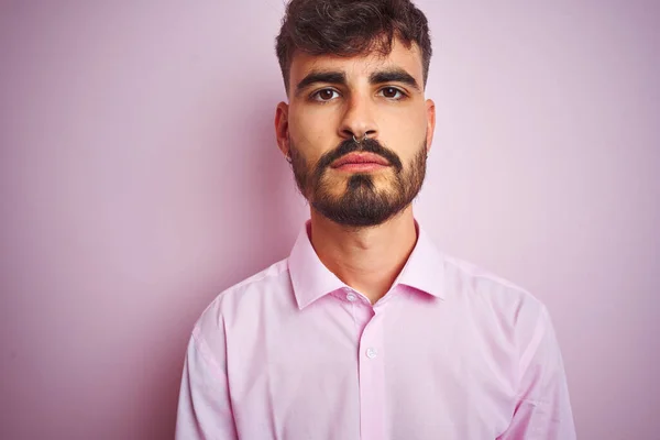 Young man with tattoo wearing shirt standing over isolated pink background with serious expression on face. Simple and natural looking at the camera.