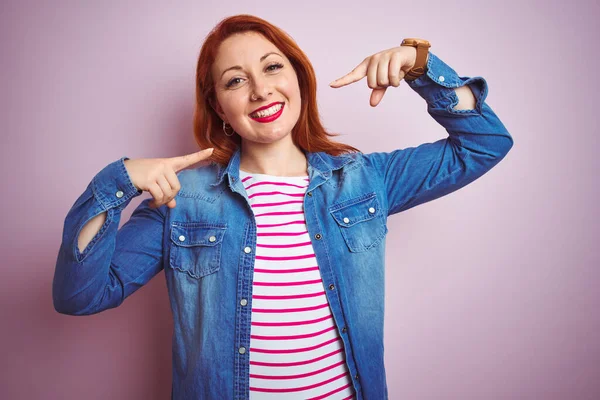 Beautiful redhead woman wearing denim shirt and striped t-shirt over isolated pink background smiling cheerful showing and pointing with fingers teeth and mouth. Dental health concept.