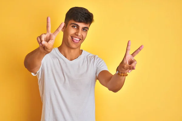 白いTシャツを着た若いインド人男性が孤立した黄色の背景の上に立っていて 両手の指が勝利サインをしているのを舌で笑っている 第二番 — ストック写真