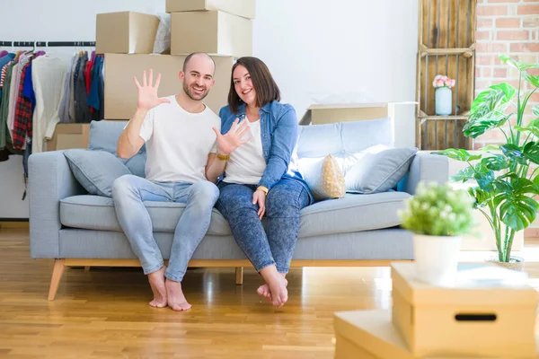 Young couple sitting on the sofa arround cardboard boxes moving to a new house showing and pointing up with fingers number ten while smiling confident and happy.