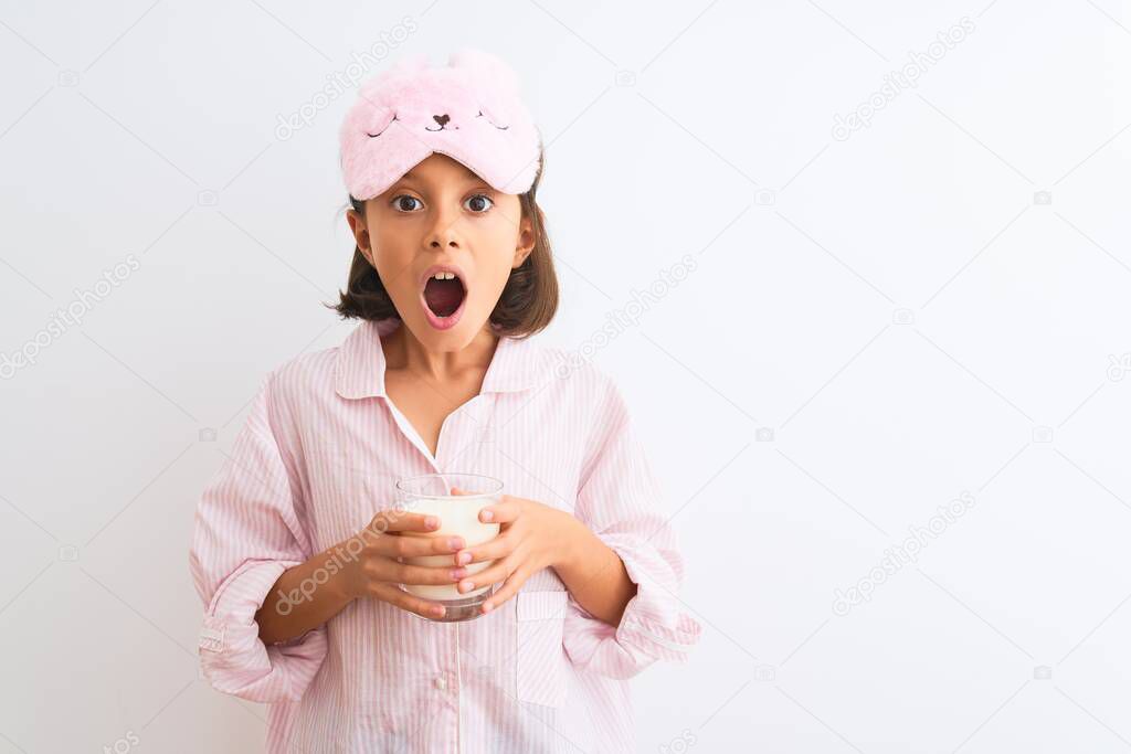 Child girl wearing sleep mask and pajama drinking glass of milk over isolated white background scared in shock with a surprise face, afraid and excited with fear expression