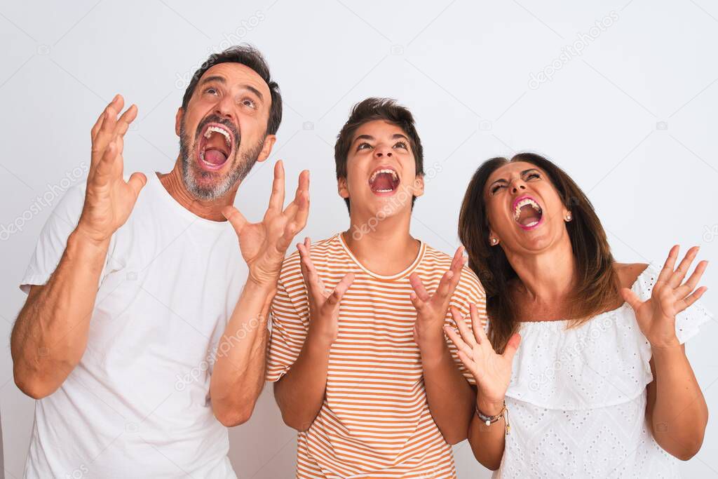 Family of three, mother, father and son standing over white isolated background crazy and mad shouting and yelling with aggressive expression and arms raised. Frustration concept.