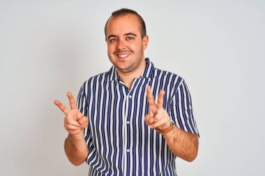 İzole edilmiş beyaz arka planda duran mavi çizgili gömlekli genç adam kameraya bakıp zafer işareti yaparken gülümsüyor. İki numara..