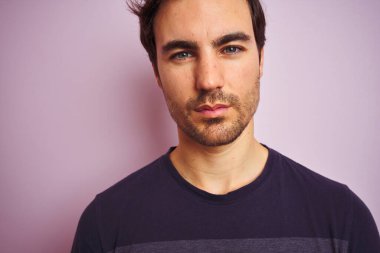 Genç yakışıklı bir adam, günlük tişört giyip pembe arka planda dikiliyor. Kendine güvenen bir yüz ifadesi var ve ciddi düşünüyor.