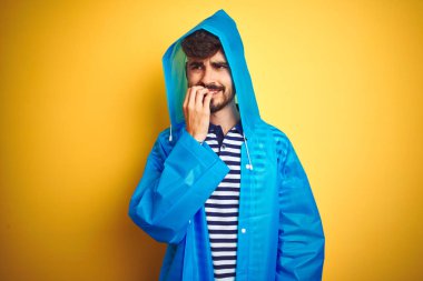 Kapüşonlu genç ve yakışıklı bir adam izole edilmiş sarı arka planda duruyor. Ağzı tırnak ısıran elleri yüzünden gergin ve stresli görünüyor. Endişe sorunu.