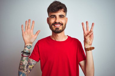 İzole edilmiş beyaz arka plan üzerinde duran kırmızı tişörtlü genç adam sekiz numaralı parmaklarıyla gülümseyerek kendinden emin ve mutlu görünüyor..