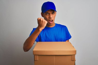 Genç yakışıklı Arap teslimatçı, izole edilmiş beyaz arka planda duran bir kutu tutuyor sinirlenmiş ve sinirlenmiş bir şekilde bağırıyor, çıldırmış ve el kaldırarak bağırıyor.