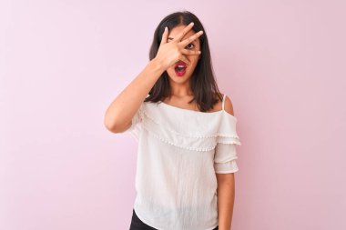 Güzel Çinli kadın, beyaz tişört giyip pembe arka planda dikiliyor. Şok olmuş bir şekilde yüzünü ve gözlerini kapatıyor. Utanmış bir ifadeyle parmaklarının arasından bakıyor..