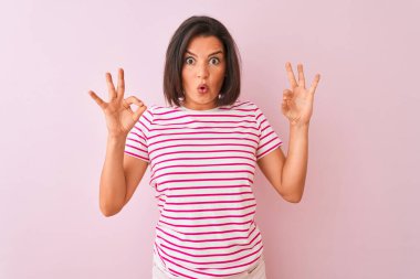 İzole edilmiş pembe arka plan üzerinde duran çizgili tişört giyen genç ve güzel bir kadın parmaklarıyla onay sembolü yaparken şaşırmış ve şok olmuş görünüyor. Çılgın ifade