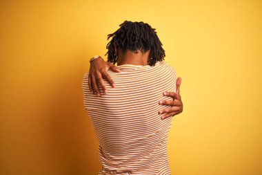 Sarı tişörtlü Afro bir adam izole edilmiş sarı arka planda dikiliyor mutlu ve pozitif bir şekilde kendine sarılıyor. Sevgi ve özgüven.