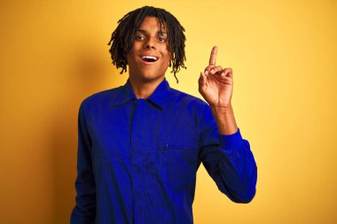 Sarı arka plan üzerinde mekanik üniforma giyen Afrikalı işçi başarılı bir fikirle parmağı işaret ediyor. Heyecanlı ve mutlu. Bir numara..