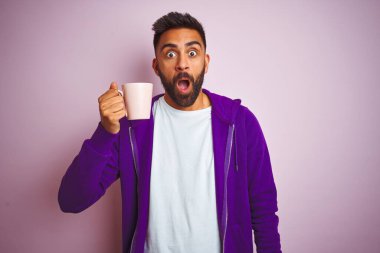 Mor kazak giyen, pembe arka planda bir fincan kahve içen Hintli adam şaşırmış bir yüzle korkmuş ve korku ifadesinden heyecanlanmış.