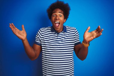 Afro saçlı, çizgili polo giyen Afrikalı bir adam, izole edilmiş mavi arka planda durmuş çılgın ve başarısını kutluyor, kollarını kaldırmış ve gözleri açık bir şekilde heyecanla haykırıyor. Kazanan konsept