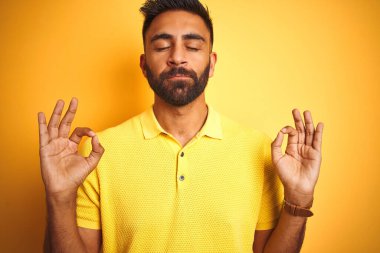 İzole edilmiş sarı arka planda polo giyen genç Hintli adam rahatla ve parmaklarıyla meditasyon hareketi yaparken gözleri kapalı gülümse. Yoga konsepti.