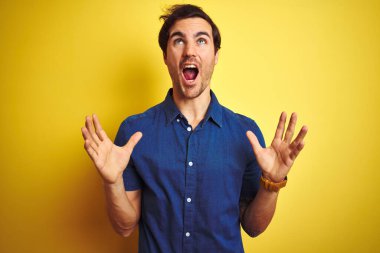 İzole edilmiş sarı arka planda dikilen sıradan tişörtlü genç yakışıklı adam sinirli bir şekilde bağırıyor ve agresif bir ifade ve kolla bağırıyor. Hayal kırıklığı konsepti.