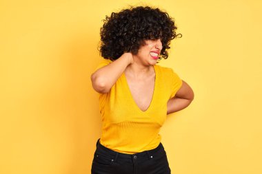 Kıvırcık saçlı genç Arap kadın izole edilmiş sarı arka planda duran tişörtü giyiyor boynu ağrıyor, eliyle boynuna dokunuyor, kasları ağrıyor.
