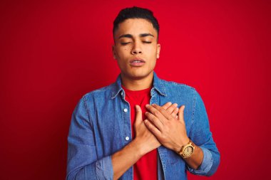 İzole edilmiş kırmızı arka plan üzerinde duran kot gömlek giyen genç Brezilyalı adam elleri göğsünde, gözleri kapalı ve yüzünde minnet dolu bir jest ile gülümsüyor. Sağlık kavramı.
