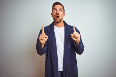 Izole arka plan üzerinde sabah rahat pijama ve bornoz giyen genç adam şaşırttı ve yukarı bakarak ve parmakları ile işaret ve kolları kaldırdı şaşırttı.
