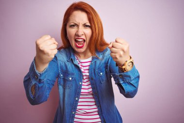 Güzel kızıl saçlı kadın kot gömlek ve çizgili tişört giyerek izole pembe arka planda kızgın ve kızgın yumruklar sallarken öfkeyle bağırıyordu. Öfke ve agresif kavram.