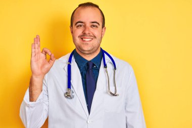 Paltolu ve steteskoplu genç doktor izole edilmiş sarı arka planda duruyor. Gülümsüyor ve el ve parmaklarıyla tamam işareti yapıyor. Başarılı ifade.