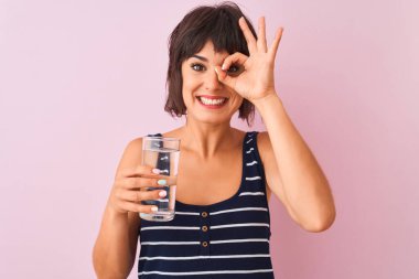 Genç, güzel bir kadın, elinde bir bardak su tutarak mutlu yüzüyle pembe arka planda duruyor.