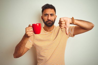 Genç Hintli adam kırmızı fincan kahve içiyor. İzole edilmiş beyaz arka planda duruyor. Kızgın bir yüzü var.
