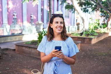 Genç ve güzel bir kadın mutlu bir şekilde gülümsüyor. Güneşli bir yaz gününde akıllı telefon kullanarak şehir sokaklarında yürüyor.